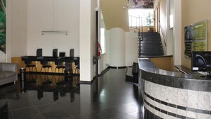 A hotel lobby with a reception desk, a staircase, and a view of a building through the window. - Hotel Primavera (Bom Despacho)