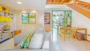 Una sala moderna con un sofá gris, una mesa de comedor de madera y sillas. Se ve una escalera con barandillas de madera, una zona de cocina con armarios verdes, y un cuadro en la pared.