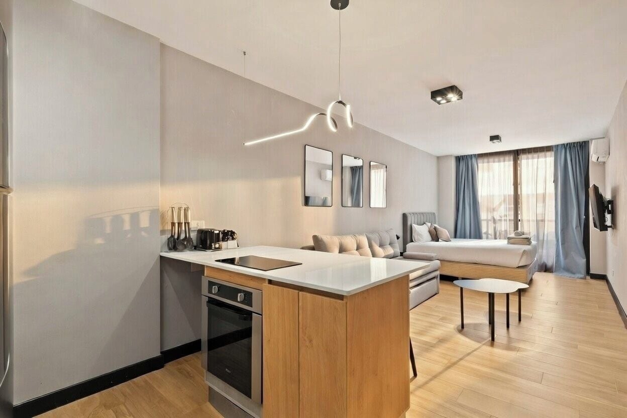 A modern kitchen with a central island, a dining area with a sofa, and a bedroom with a bed and curtains.