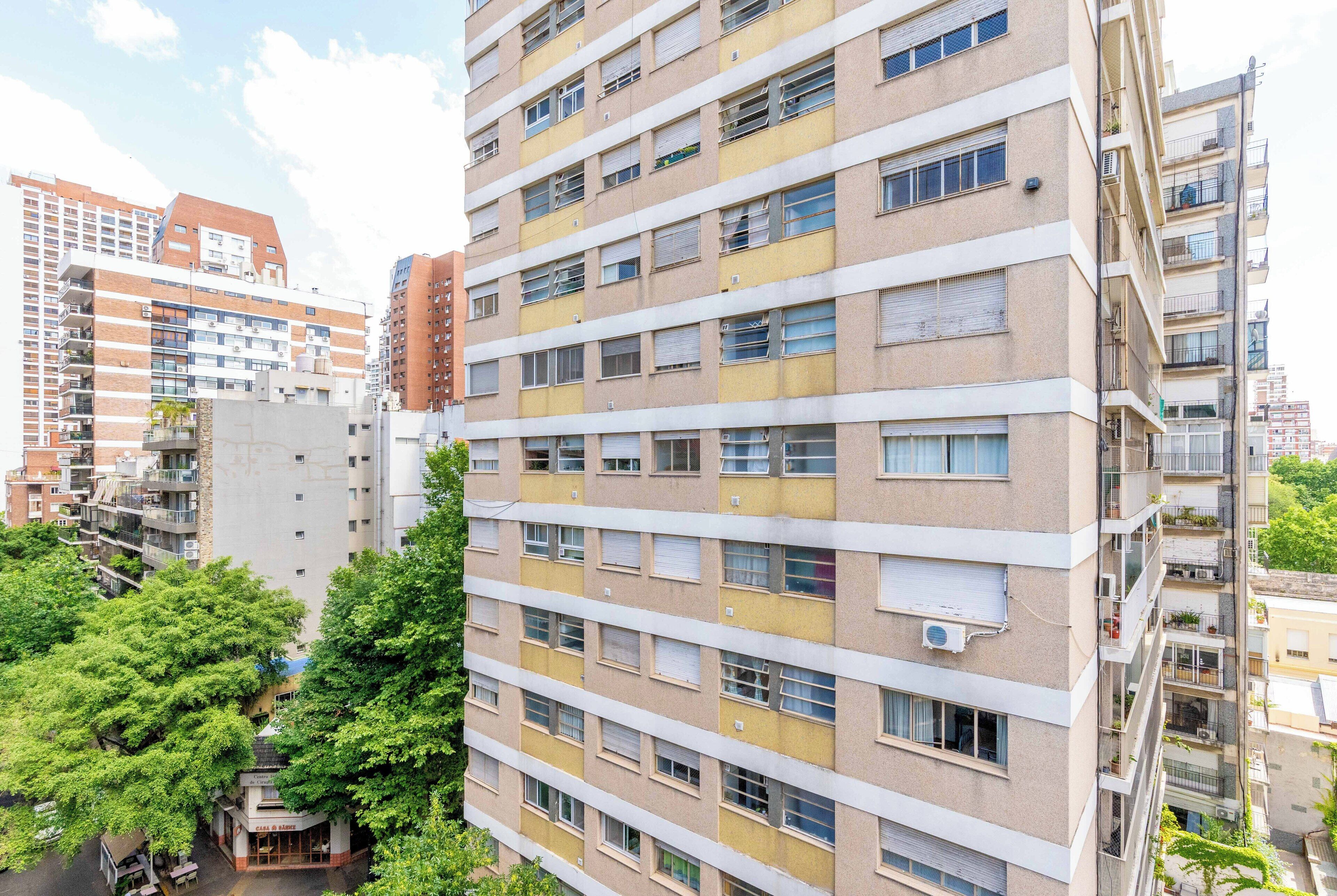 A multi-story residential building with balconies, a ground-level entrance, and surrounding greenery.