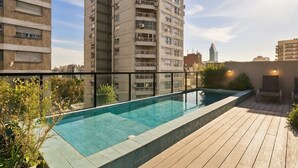 Ein Pool auf dem Dach mit einer Holzterrasse, umgeben von hohen Gebäuden, und einem wolkenlosen Himmel.