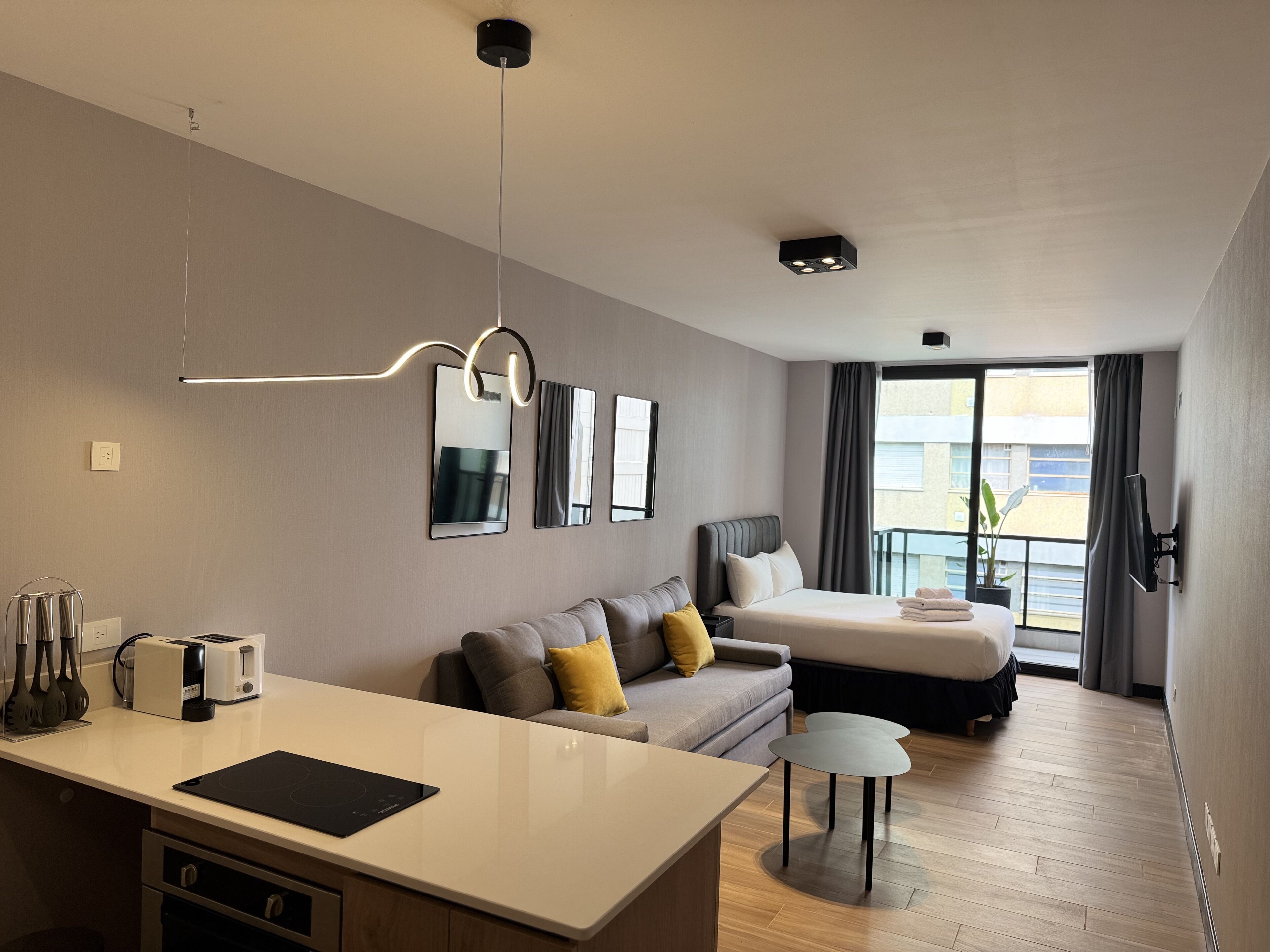 A modern hotel room with a kitchenette, a sofa, a bed, and a balcony with a view.