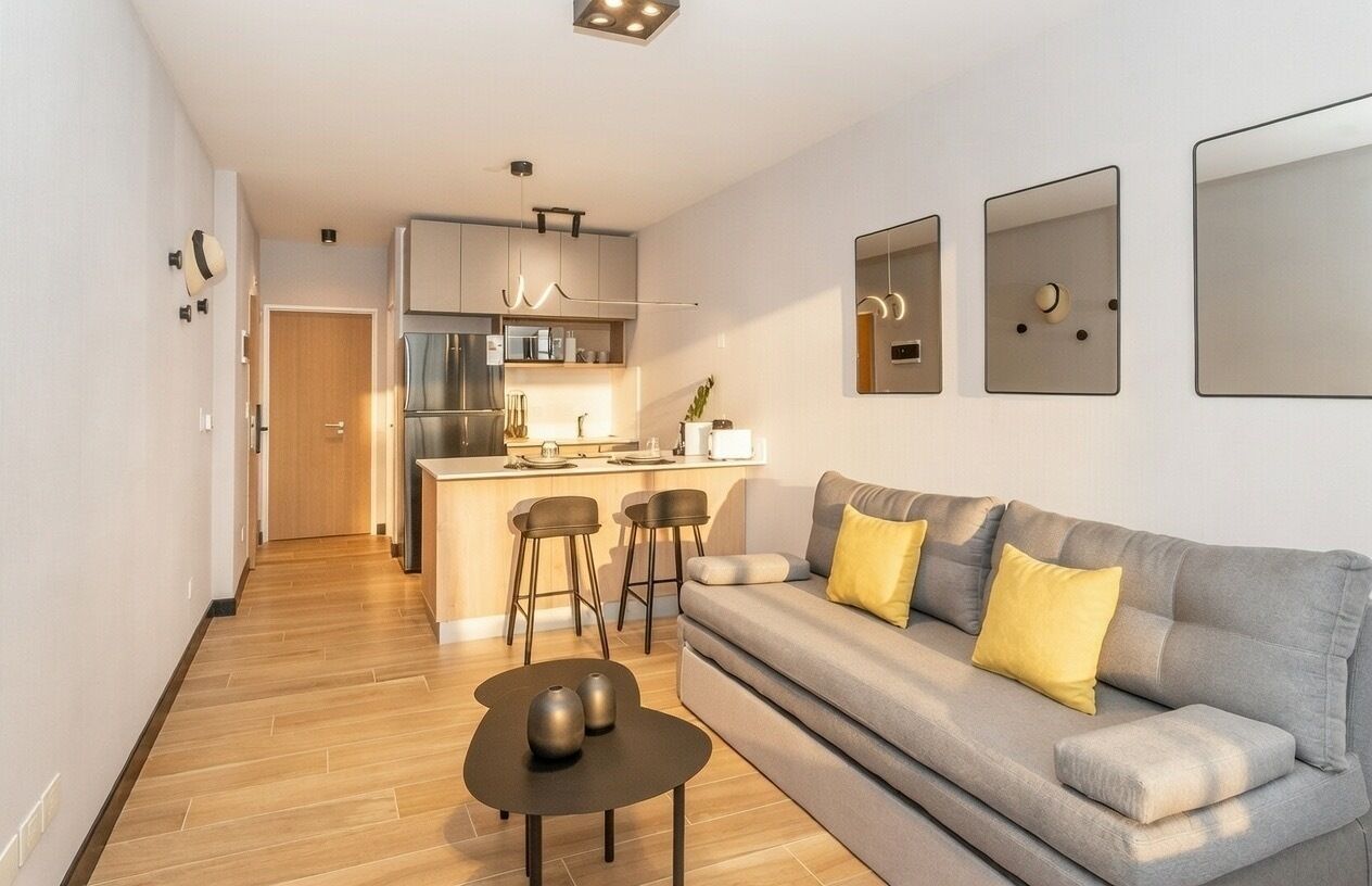 Una sala de estar moderna con un sofá gris, cojines amarillos, una mesita negra y una cocina al fondo.