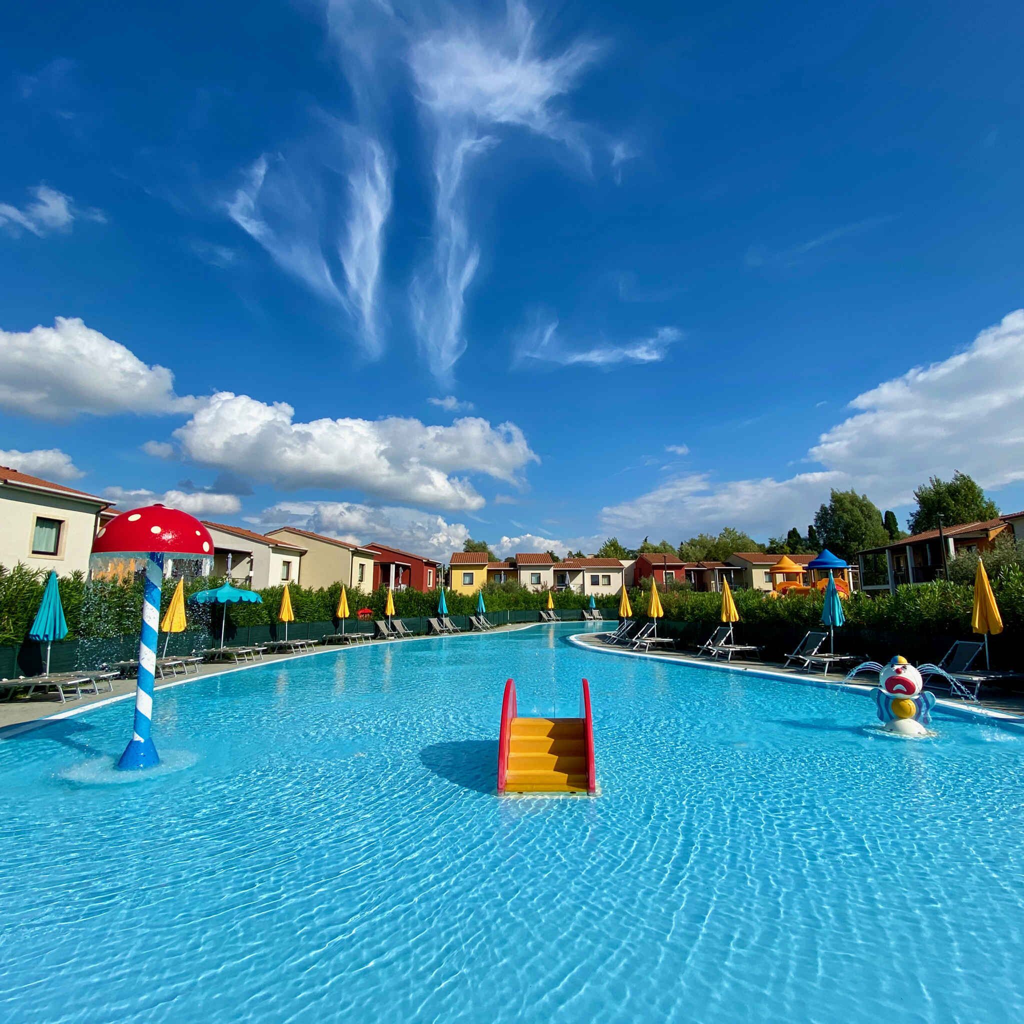 Une piscine avec un toboggan, des chaises longues, des parasols et un ciel d’un bleu éclatant.