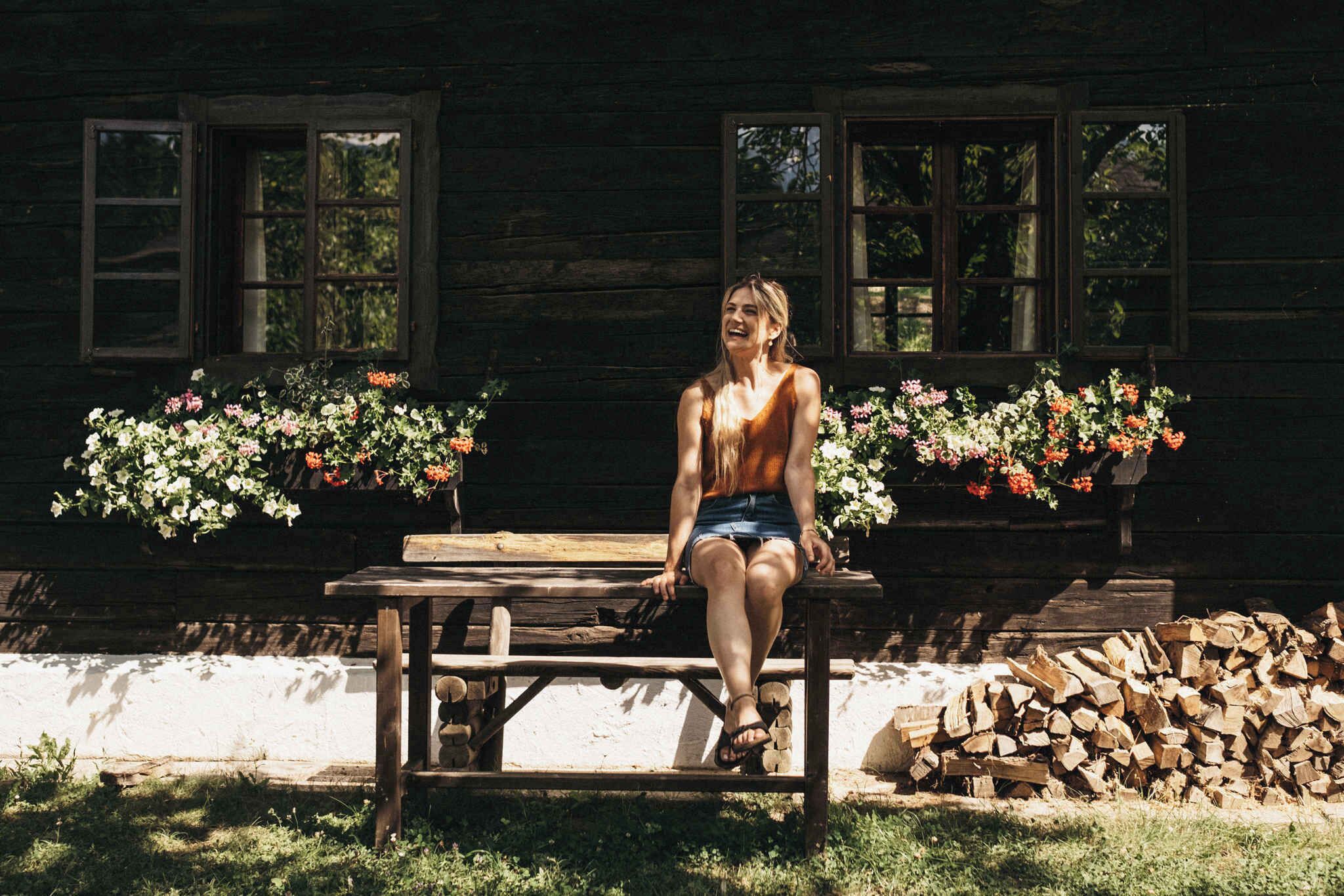 Une personne est assise sur un banc en bois devant un bâtiment rustique également en bois, dont les rebords de fenêtres sont ornés de jardinières.