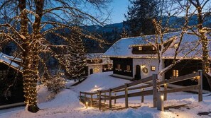 Eine verschneite Landschaft mit einem Haus, das mit Lichterketten geschmückt ist, einem Holzzaun und einem mit Schnee bedeckten Hügel.