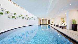 Une piscine intérieure moderne, avec une eau d’un bleu limpide, entourée de chaises longues et de plantes en pot disposées le long des murs.