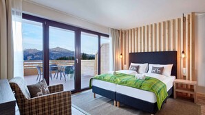 Une chambre avec un grand lit, une chaise, une table de chevet et une vue sur les montagnes, visible à travers une porte coulissante.
