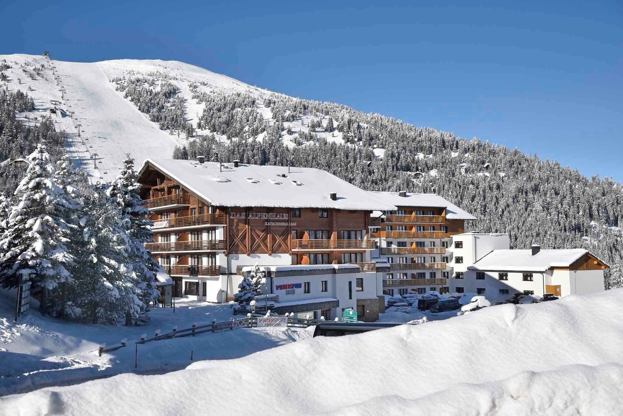 Estância de esqui nas montanhas coberta de neve, com vários edifícios incluindo uma hospedaria e um hotel, cercada por árvores e uma pista de esqui.