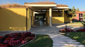 A yellow building with a covered entrance, a pathway leading to the entrance, and flower beds with pink and red flowers. - Holiday apartment with pool (Castelnuovo del Garda)