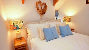 A bedroom with a bed, pillows, a nightstand, a wall-mounted light, and a decorative wreath. - Porthcothan Mill Cottage (Porthcothan Bay)