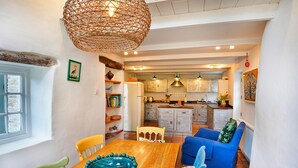 A dining area with a wooden table, chairs, and a decorative bowl. A kitchen is visible in the background with cabinets and a hanging light fixture. - Porthcothan Mill Cottage (Porthcothan Bay)