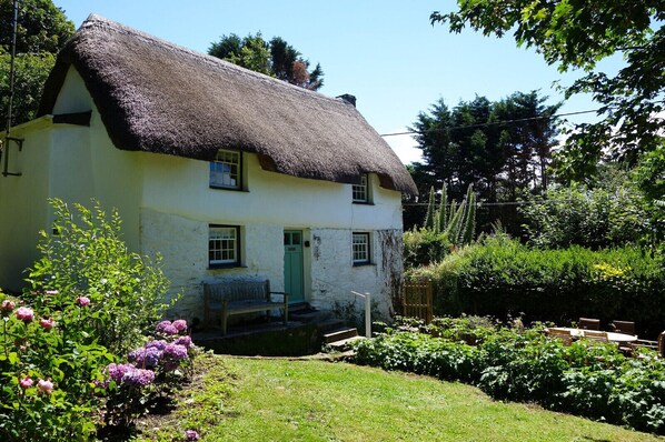 A traditional thatched-roof house with a green door, surrounded by lush greenery and flowering plants.