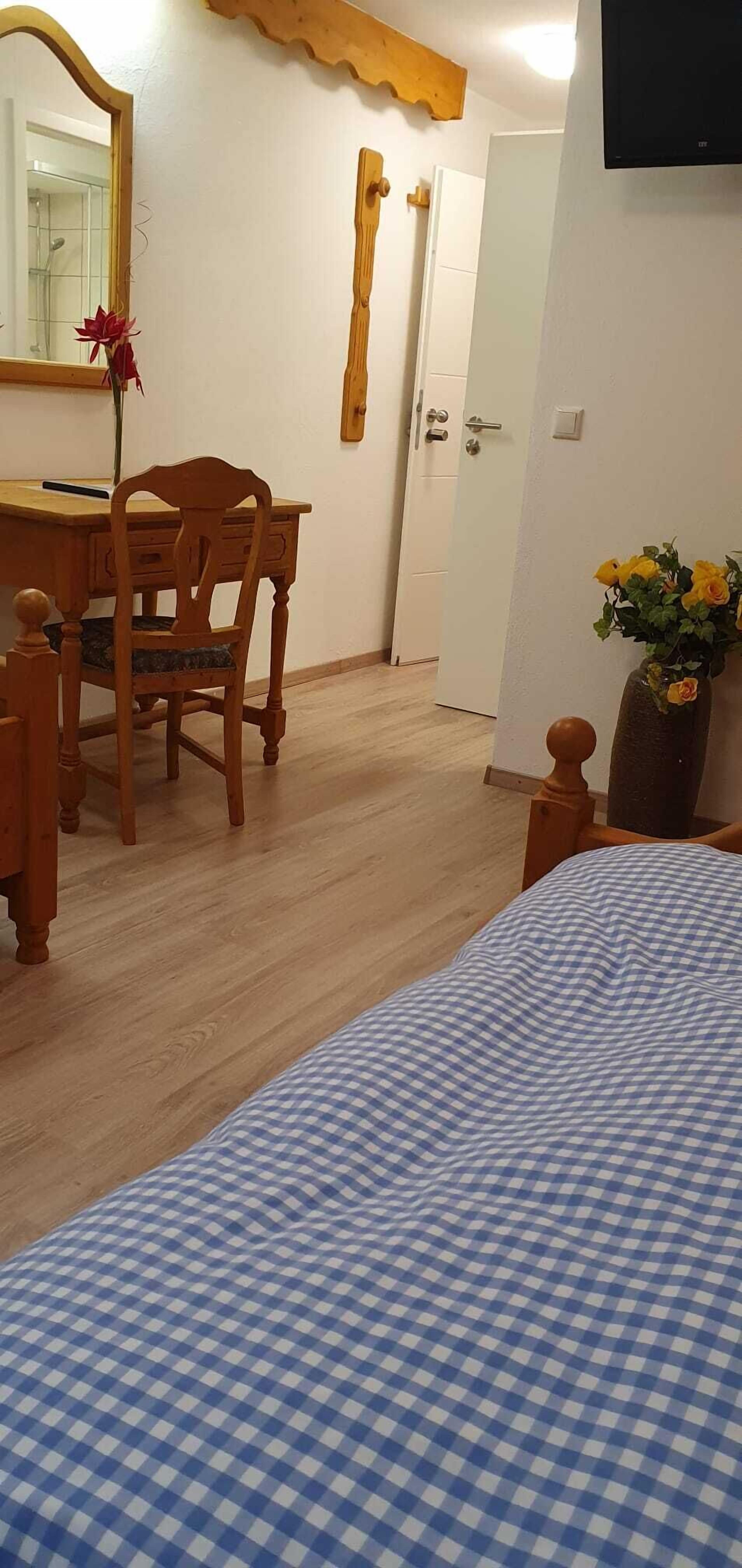 A bedroom with a checkered bedspread, a wooden desk with a chair, a mirror, and a vase with yellow flowers.