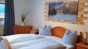 A hotel room with a bed, a nightstand with a lamp, a dresser, a vase with branches, and a framed picture of a lake. - Berggasthof Willy Merkl (Schliersee)