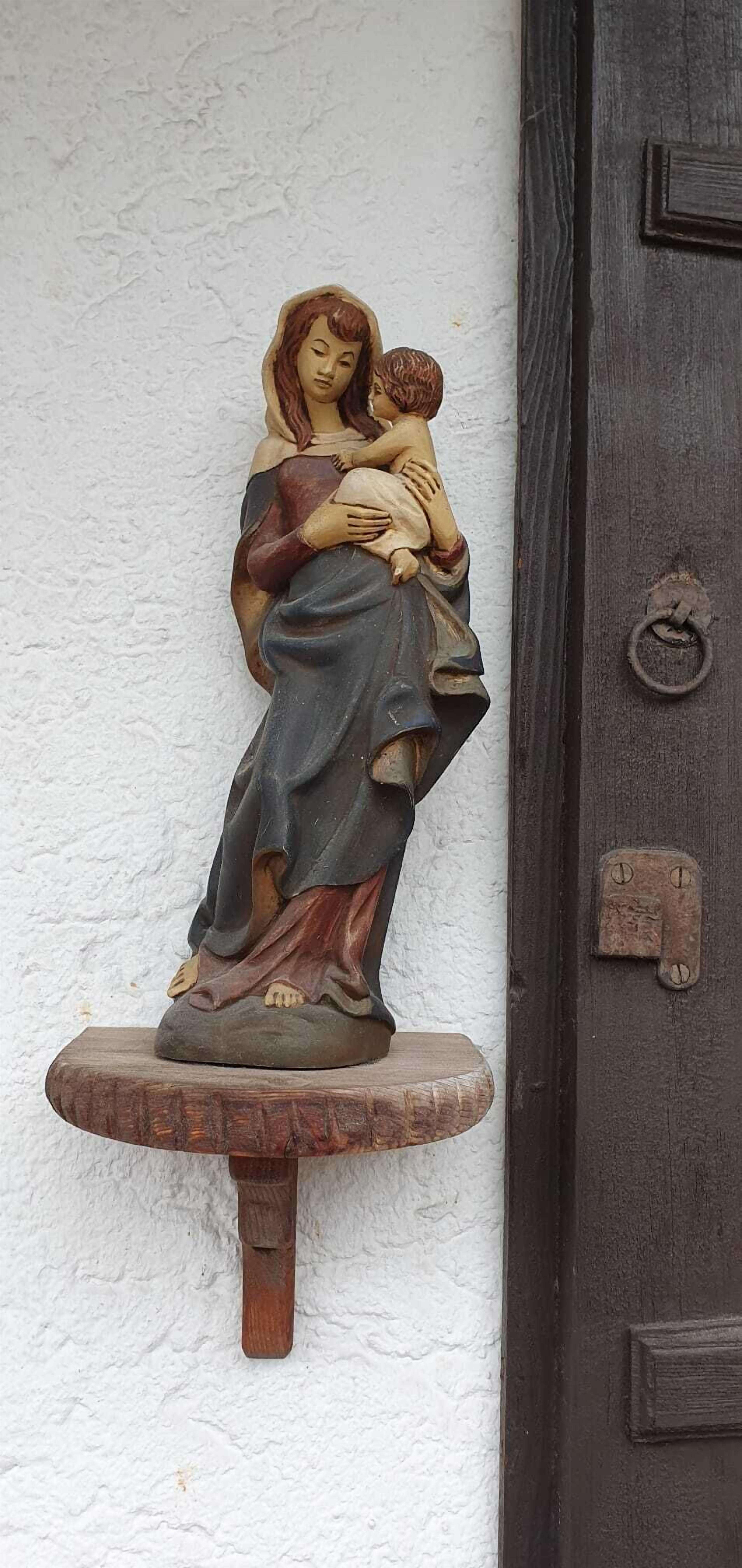 Eine hölzerne Statue einer Frau, die ein Kind hält, auf einem Holzfußsockel angebracht, an einer weißen Wand neben einer dunklen Holztür mit einem Metallring als Griff.