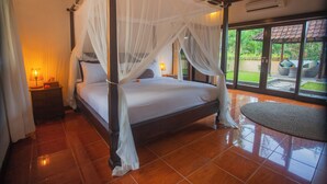 A four-poster bed with a canopy, a nightstand with a lamp, and a view of the outdoors through large windows. - Coco Villa (Ubud)