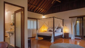 A four-poster bed with a canopy, a bedside table with a lamp, a bathroom with a sink and mirror, and a wooden ceiling. - Coco Villa (Ubud)