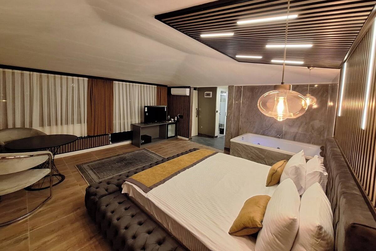 A modern hotel room with a large bed, a seating area with a sofa and chairs, a desk with a flat-screen TV, a bathroom with a bathtub, and a ceiling with linear lighting fixtures.