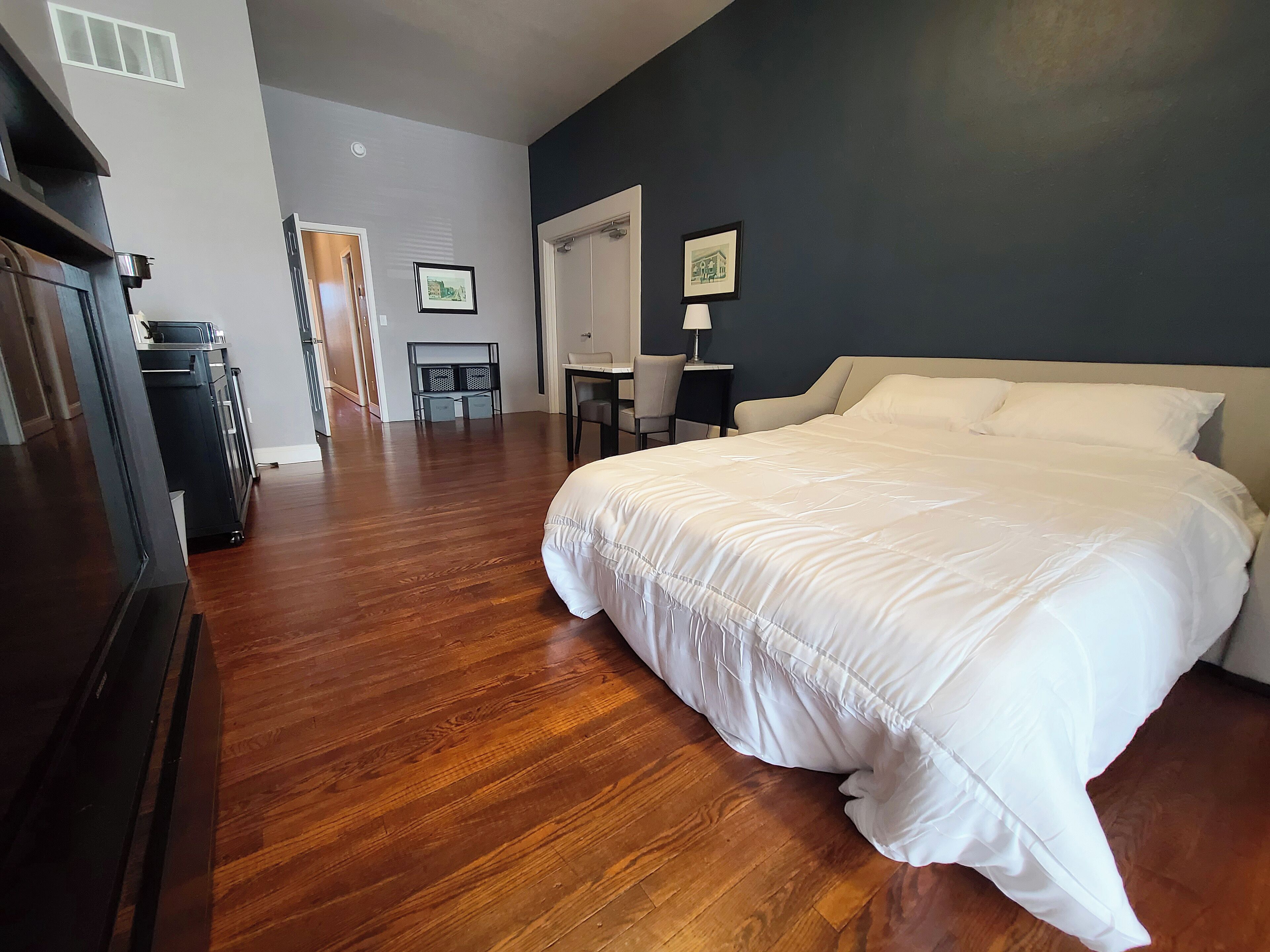 A bedroom with a large bed, wooden flooring, a desk, and a chair.