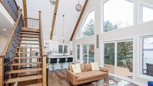 A spacious living area with a wooden staircase, a kitchen with white cabinets, a large window overlooking a deck, and a glass coffee table.