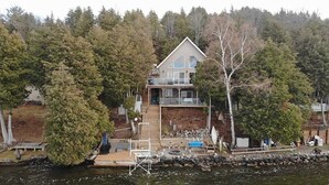 A two-story house with a deck, surrounded by dense forest and a body of water.