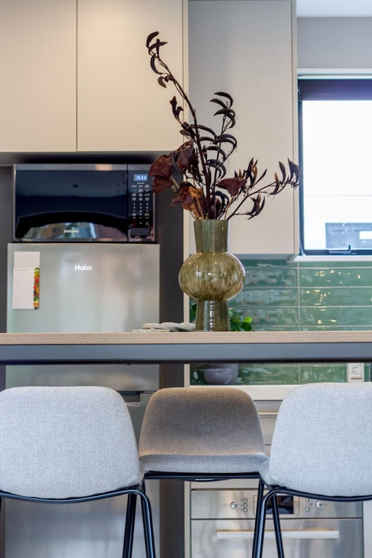 Eine moderne Küche mit Mikrowelle, einem Haier-Kühlschrank und einer Vase mit getrockneten Blumen auf der Arbeitsplatte. Vor der Arbeitsplatte stehen zwei graue Stühle.