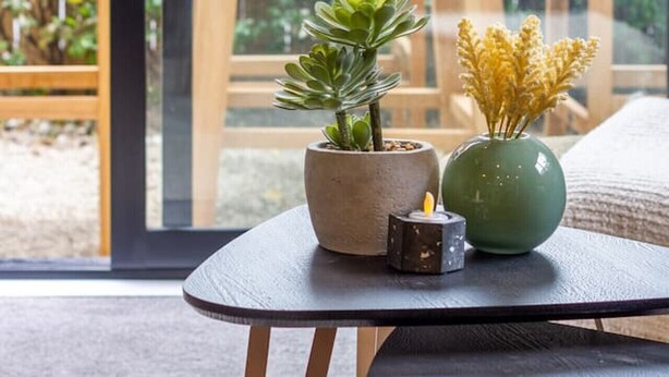 Uma pequena mesa com duas plantas em vasos e uma vela, colocada em frente a uma grande janela com vista para um deck de madeira e vegetação.