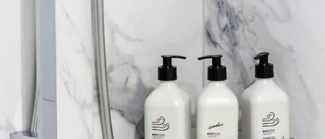 A bathroom with a shower, a glass shelf holding three bottles, and a marble wall.