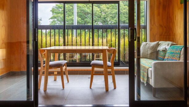 Une pièce dotée d’une porte-fenêtre coulissante donnant sur un balcon avec une table et des chaises en bois, un canapé et une vue sur la verdure.