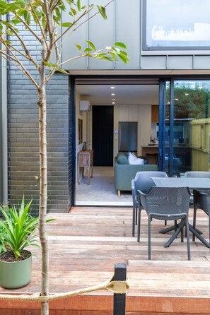 A modern outdoor patio area with a dining table and chairs, a small potted plant, and a view into a contemporary interior space. - Merivale Gem: 2BR Townhouse Oasis (Christchurch)