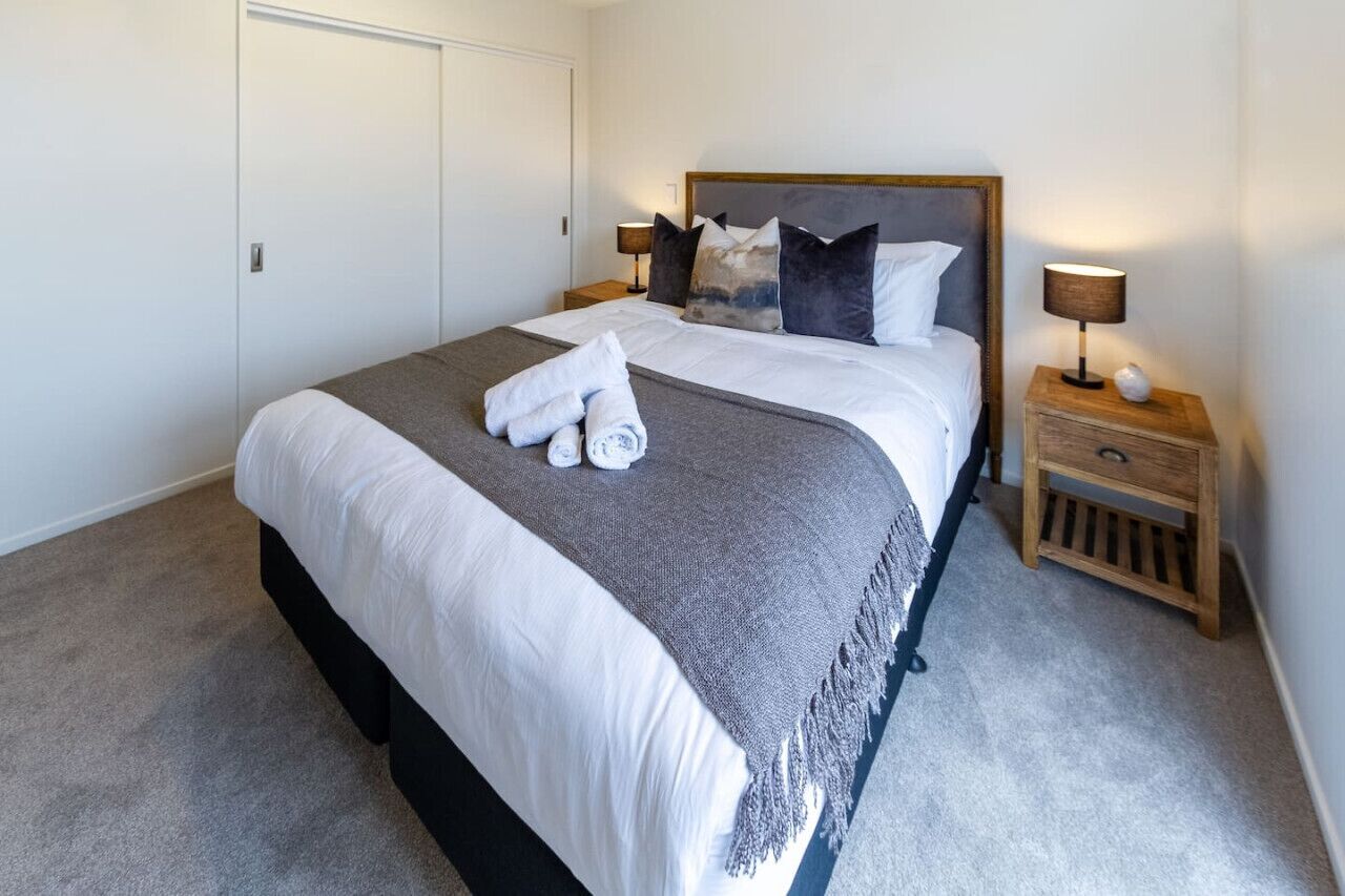 A neatly made bed with a grey blanket and rolled towels, a wooden nightstand with a lamp, and a closed door in a hotel room.