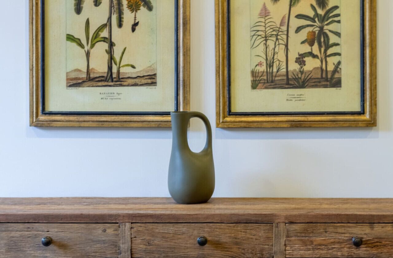 Une illustration botanique encadrée orne un mur, et un vase est posé sur une commode en bois en dessous.