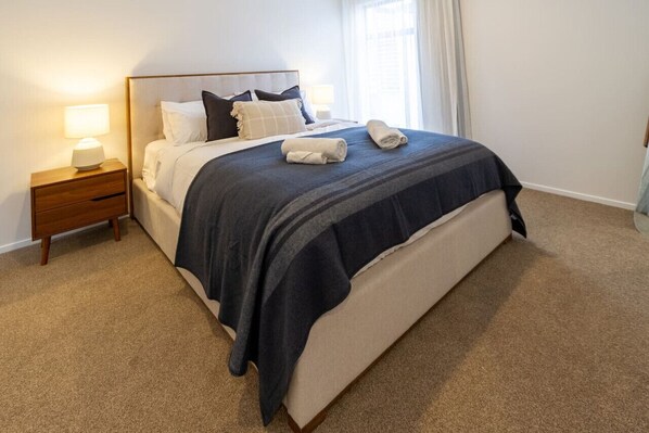 A neatly made bed with a dark blue bedspread and white pillows, a wooden nightstand with a lamp, and a window with curtains.