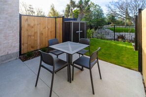 Un patio con una mesa y cuatro sillas, cerca de madera, un cobertizo y un césped bien cuidado.