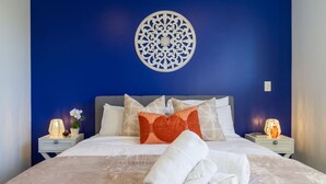 A neatly made bed with white linens and a heart-shaped pillow, two bedside lamps, and a decorative wall piece.