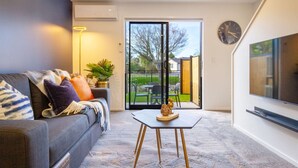 A living room with a grey sofa, a small table with a decorative item, a TV on the wall, and a sliding glass door leading to a garden.