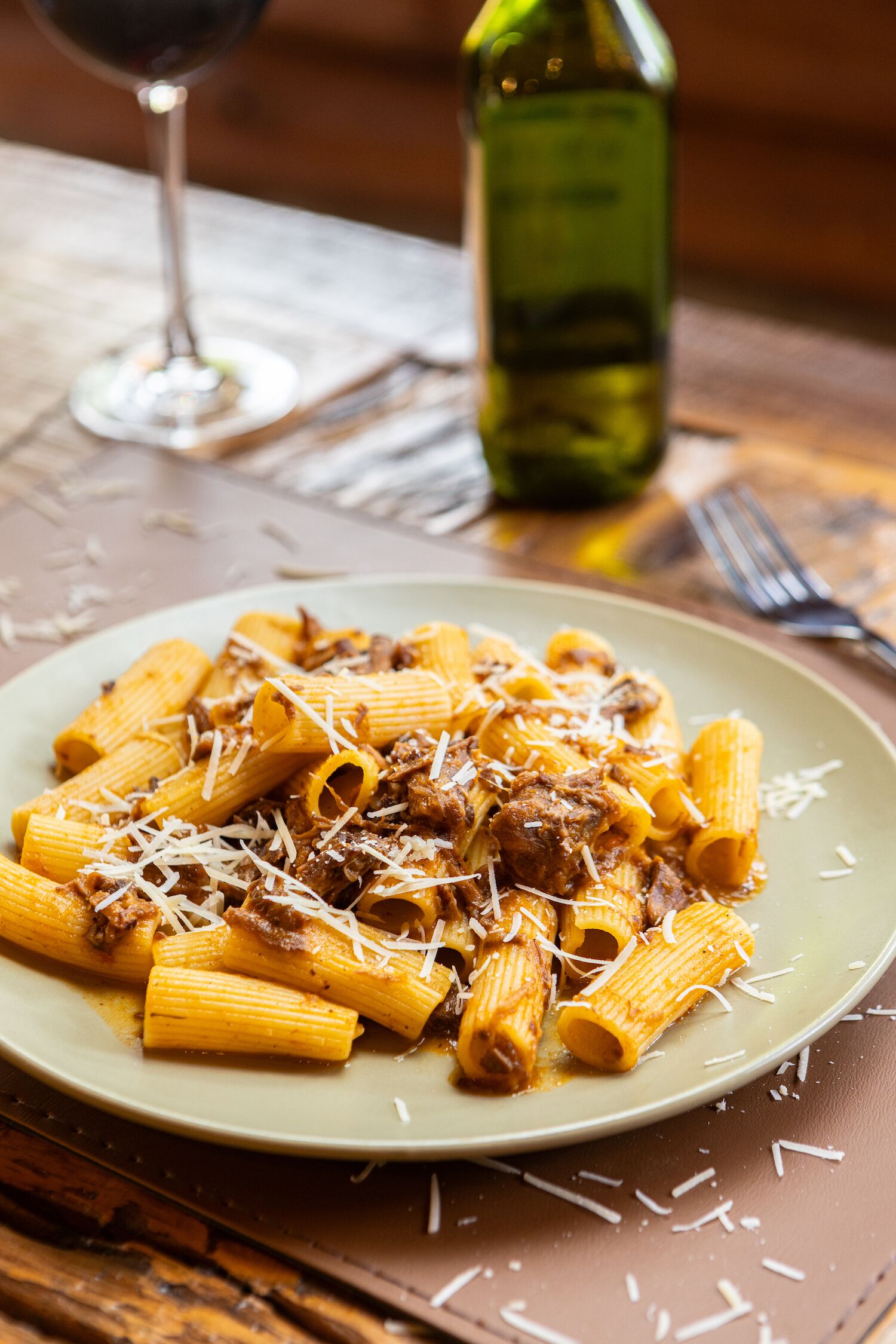 A plate of pasta with meat sauce and grated cheese, a wine glass, and a bottle of wine on a wooden table.