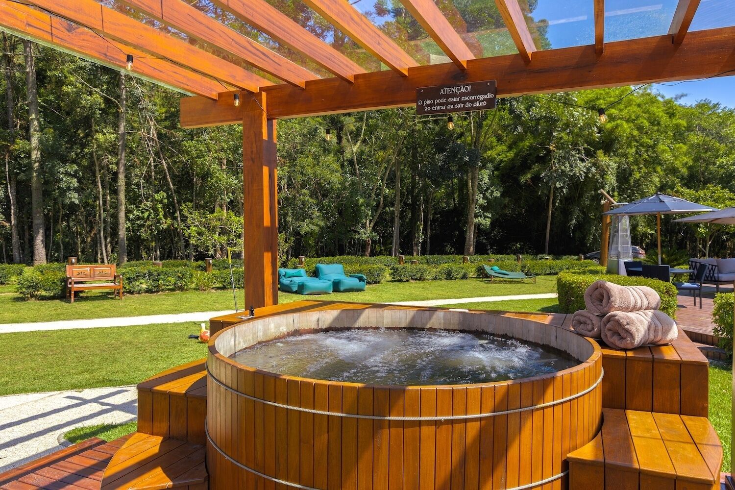 A wooden hot tub with a wooden pergola overhead, surrounded by greenery and outdoor seating.