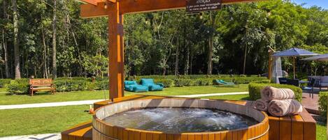 A wooden hot tub with a wooden pergola overhead, surrounded by greenery and outdoor seating.