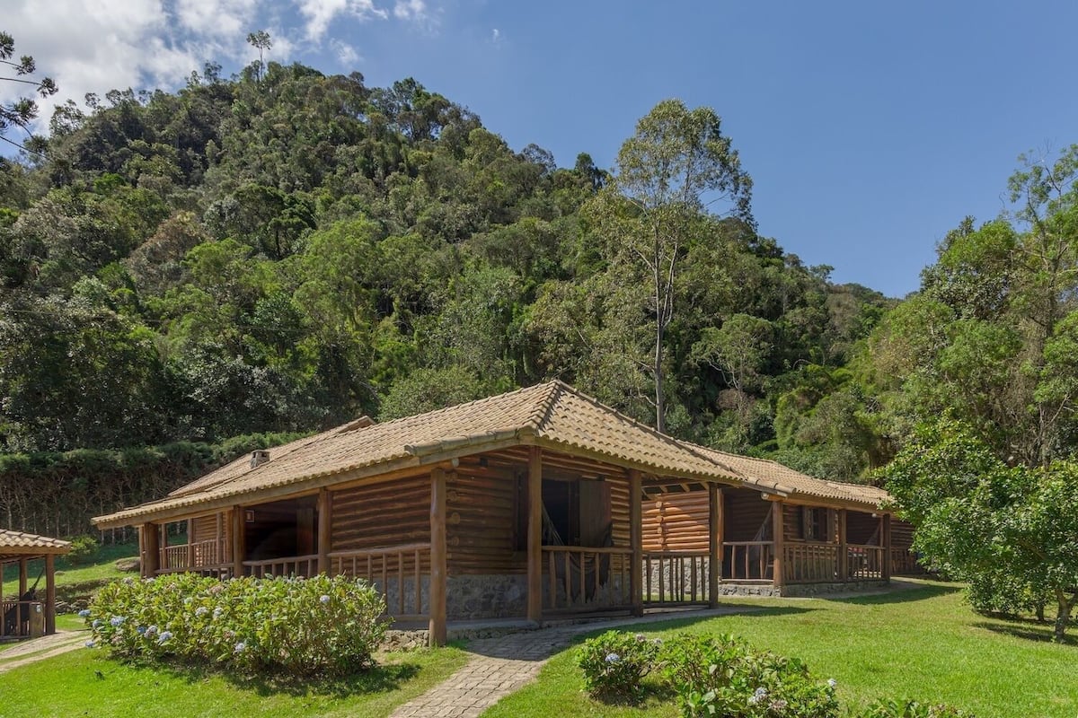 A wooden cabin with a tiled roof, surrounded by lush greenery and a mountainous backdrop.