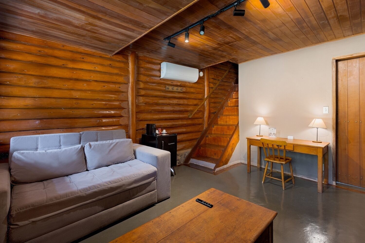 Interior de una cabaña de troncos con un sofá, una mesita, un escritorio con dos lámparas y una escalera.