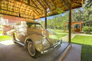 Une voiture ancienne est garée à l’intérieur d’une structure abritée, d’où l’on peut admirer un jardin et des arbres.