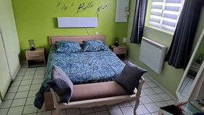 A bedroom with a bed, nightstands, a bench, a wall with a decorative sign, a window with curtains, and a radiator.