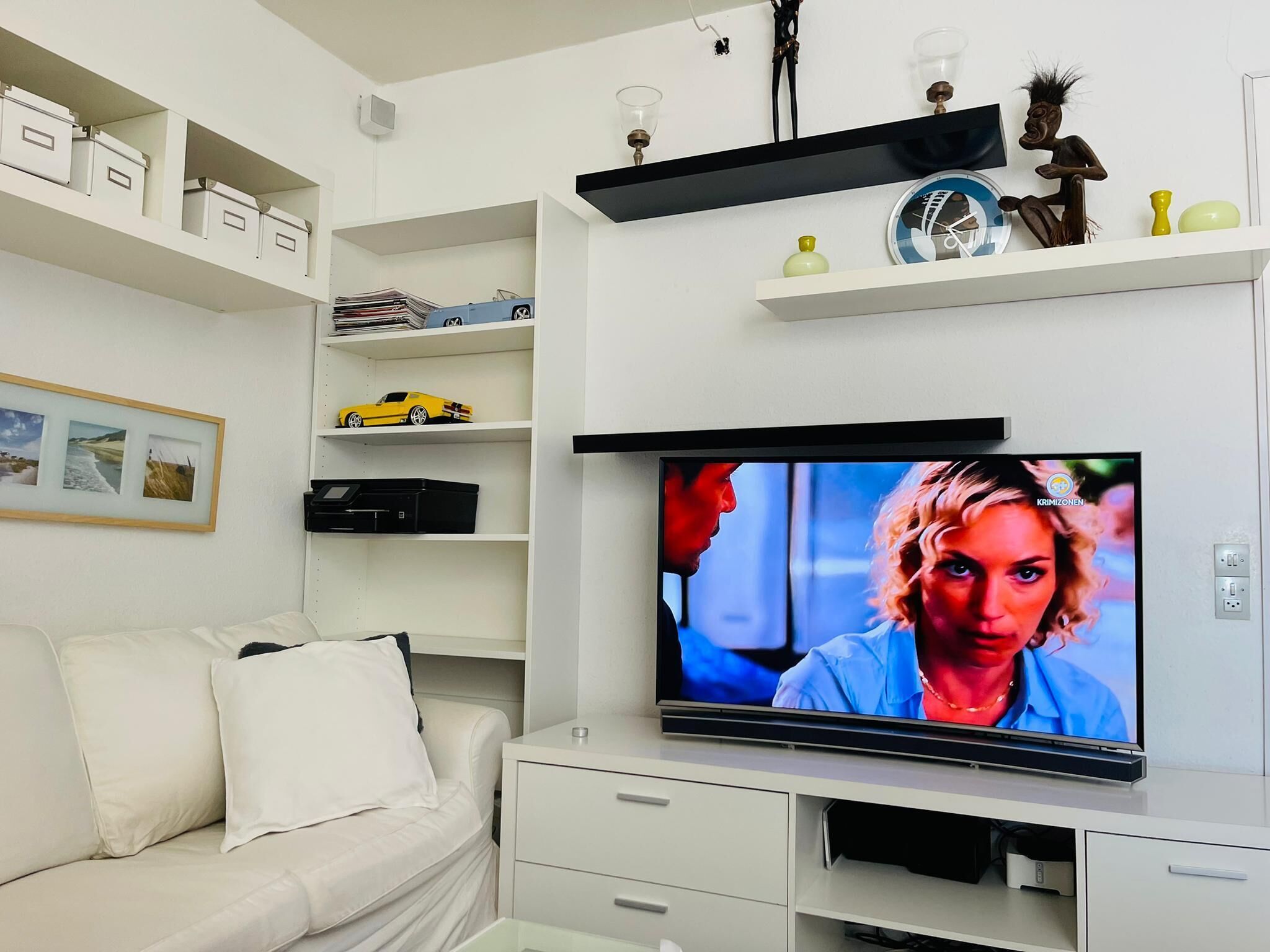 A living room with a flat-screen TV mounted on the wall, a white sofa, and shelves with various items including a yellow toy car and decorative objects.