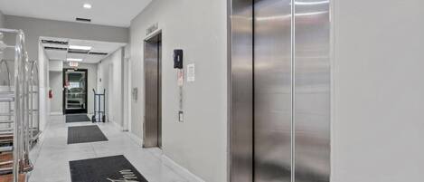 A hotel corridor with a clean, modern design, featuring a central elevator and a reception area visible in the distance.