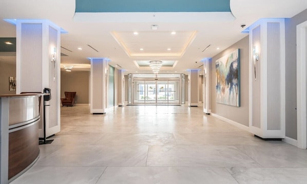 A spacious hotel corridor with a modern design, featuring a central light fixture, glass doors leading to a room, and a reception area with a counter and chairs. - Ocean Side Inn Studios (Daytona Beach Shores)