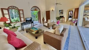 A spacious living area with a sofa, armchairs, a coffee table, and a television. There are ceiling fans, a large window with a view of greenery, and a door leading to another room. - Private 3bdr ocean view villa (Cole Bay)