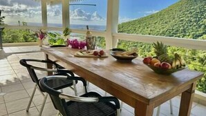 A wooden table with chairs, a bowl of fruit, and a view of a lush green hillside. - Private 3bdr ocean view villa (Cole Bay)