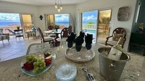 A spacious living area with a large glass door leading to a balcony with a view of the ocean. There is a dining table with chairs, a coffee table with a fruit platter, and a wine bottle on the counter. - Pelican Pearl Villa - GENERATOR (Sint Maarten)